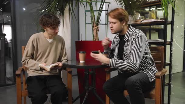 Dois gays num encontro estão sentados num café. Um tipo está a ler um livro e o outro está a mostrar alguma coisa no telemóvel. LGBT — Vídeo de Stock