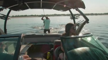 Bir kadın motorlu tekne kullanıyor, ve arka planda bir adam wakeboard kullanıyor, ve erkek ona yardım ediyor.