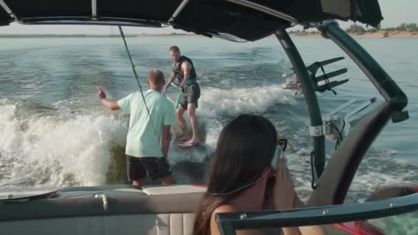 Μια γυναίκα οδηγεί ένα μηχανοκίνητο σκάφος, και στο βάθος ένας τύπος είναι καβάλα σε ένα wakeboard, και ο άνθρωπος τον βοηθά — Αρχείο Βίντεο