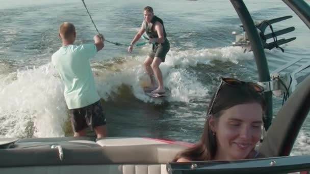 Una mujer está conduciendo un barco a motor, y en el fondo un tipo está montando un wakeboard, y el hombre lo está ayudando — Vídeo de stock