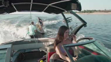 Bir kadın motorlu tekne kullanıyor, ve arka planda bir adam wakeboard kullanıyor, ve erkek ona yardım ediyor.