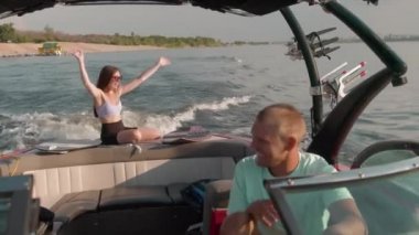 Bir adam motorlu tekne kullanıyor, ve arka planda bir kadın oturuyor ve hoş havanın tadını çıkarıyor.