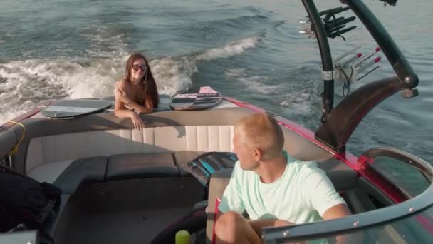 Мужчина водит моторную лодку, а женщина на заднем плане лежит на животе и наслаждается приятной погодой — стоковое видео