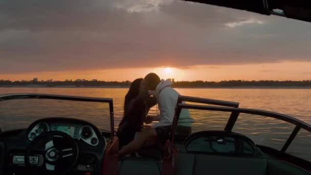 Egy fiú és egy lány ülnek egy motorcsónakban, csókolóznak és nézik a naplementét. Romantikus hangulat. — Stock videók