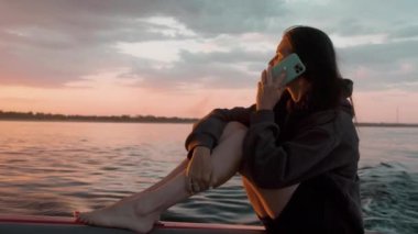 Bir kadın gün batımında motorlu bir teknede telefonla konuşuyor.