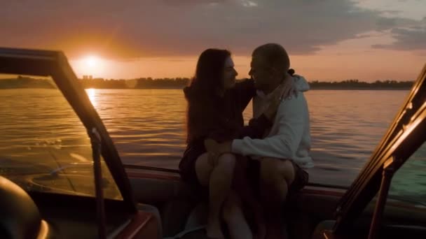 Facet i dziewczyna przytulają się słodko o zachodzie słońca siedząc na łodzi — Wideo stockowe