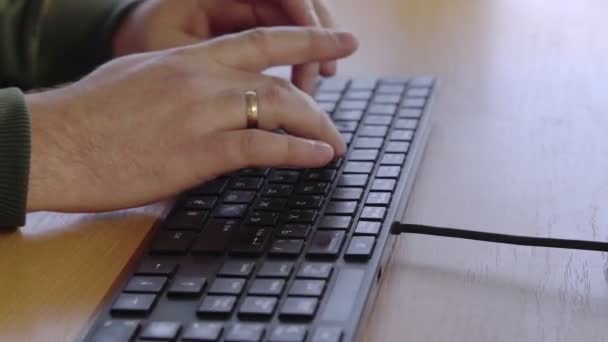 Close-up de mãos masculinas digitando em um teclado preto enquanto sentado em uma mesa dentro de casa. Vista lateral de um homem trabalhando em um computador. — Vídeo de Stock