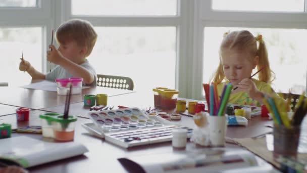 Gyerekek ülnek az asztalnál és rajzolnak különböző színekkel és ecsetekkel. — Stock videók