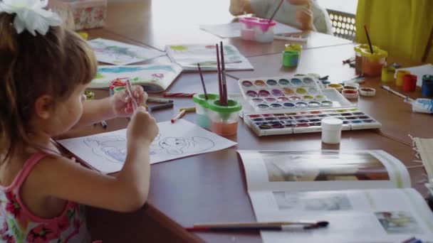 Πλευρά άποψη ένα μικρό κορίτσι κάθεται σε ένα τραπέζι και σχέδιο σε χαρτί με διαφορετικά χρώματα και πινέλα — Αρχείο Βίντεο