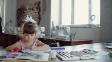 Küçük bir kız masada oturuyor ve farklı renkler ve fırçalarla kağıda resim çiziyor.