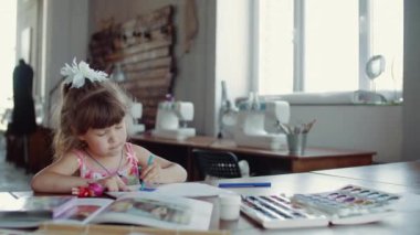 Küçük bir kız masada oturuyor ve farklı renkler ve fırçalarla kağıda resim çiziyor.