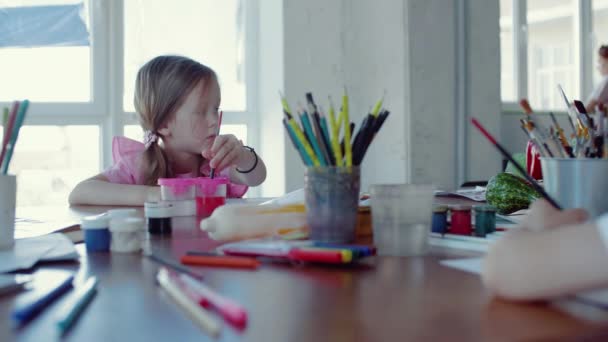 Маленькая девочка сидит за столом и рисует на бумаге разными цветами и кисточками — стоковое видео