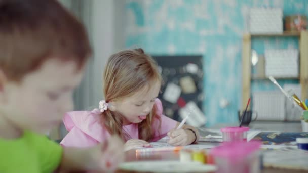 Vista laterale una bambina è seduta a un tavolo e disegna su carta con colori e pennelli diversi — Video Stock