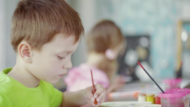 Vista lateral um menino e uma menina estão sentados a uma mesa e desenhando em papel com cores e pincéis diferentes — Vídeo de Stock