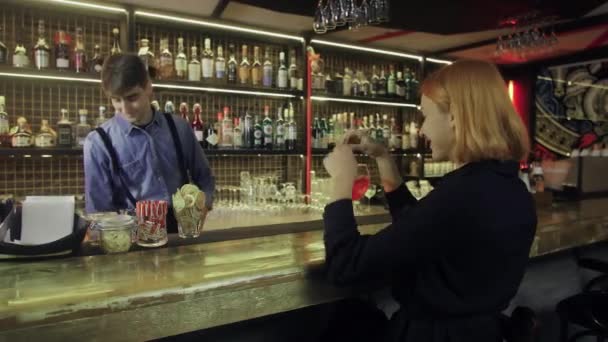 Девушка сидит в баре с коктейлем в руках и общается с барменом — стоковое видео