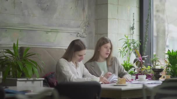 Twee meisjes zitten aan een tafel in een café en lossen zakelijke problemen op. Werken buiten het huis — Stockvideo