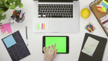 Üst manzara. Tasarımcı bir kız krom ekranda tabletle masada oturuyor. Masada çeşitli ofis malzemeleri var.