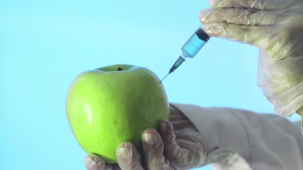 Primer plano de un humano con una capa médica y guantes inyectando una jeringa en una manzana con un poco de líquido sobre un fondo azul — Vídeos de Stock