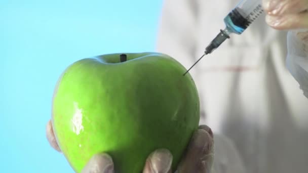 Nahaufnahme eines Menschen in medizinischem Mantel und Handschuhen, der eine Spritze in einen Apfel mit etwas Flüssigkeit auf blauem Hintergrund injiziert — Stockvideo