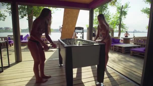 Женщины играют в настольный футбол или кикер с миниатюрными игроками на пляже в купальниках — стоковое видео