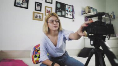 Gözlüklü genç bir kadın kanepede oturuyor gülümsüyor, kamerayla konuşuyor. İzleyicilerinizi selamlıyorum.