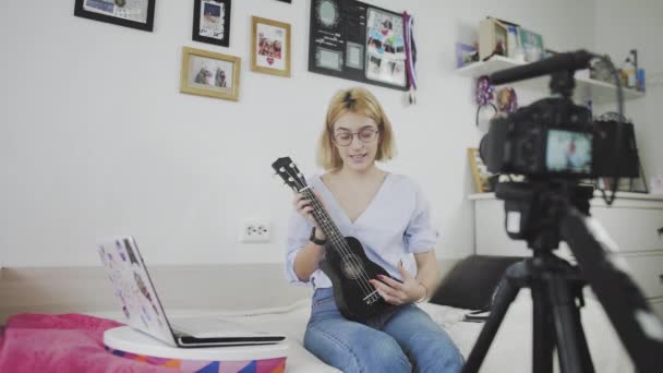 Młoda kobieta w okularach siedzi na kanapie, uśmiecha się i gra na ukulele do kamery. Gram na ukulele. Blogger wideo — Wideo stockowe
