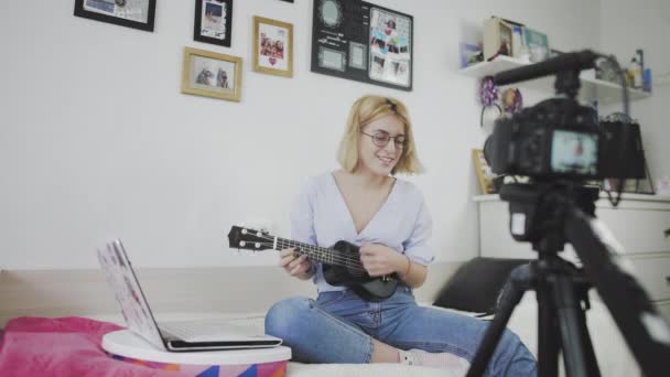 Μια νεαρή γυναίκα με γυαλιά κάθεται στον καναπέ, χαμογελώντας και παίζοντας το γιουκαλίλι στην κάμερα. Παίζω γιουκαλίλι. Blogger βίντεο — Αρχείο Βίντεο