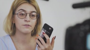 Gözlüklü, elinde telefonla kameraya gülümseyen ve konuşan genç bir kadının yakın çekimi.