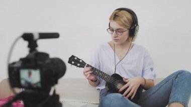 Gözlüklü genç bir kadın kanepede oturuyor, gülümsüyor ve kameraya ukulele çalıyor. Ukulele çalıyorum. Video Blogçusu