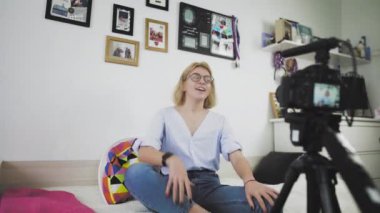 Gözlüklü genç bir kadın kanepede oturuyor gülümsüyor, kamerayla konuşuyor.