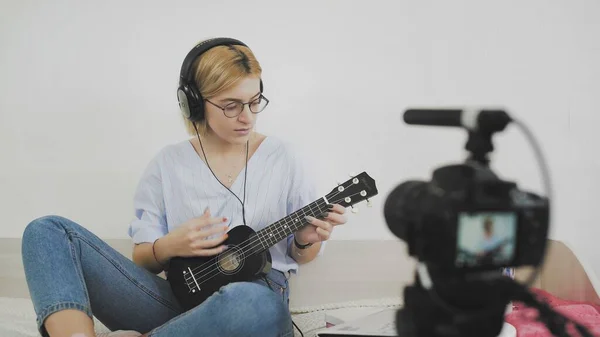 Uma jovem mulher com óculos está sentada no sofá, sorrindo e tocando o ukulele para a câmera. A tocar ukulele. Vídeo Blogger — Fotografia de Stock