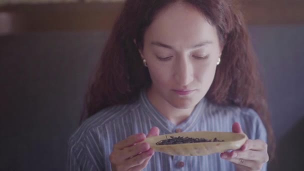 Una bella ragazza dai capelli ricci raccoglie una ciotola di legno con foglie di tè e annusa — Video Stock