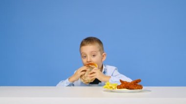 Çocuk masaya oturur, fast food yer ve mavi arka planda sevinir. Sağlıklı ve sağlıksız gıda kavramı. Fast food