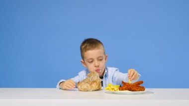 Çocuk masaya oturur, fast food yer ve mavi arka planda sevinir. Sağlıklı ve sağlıksız gıda kavramı. Fast food