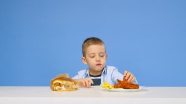 Çocuk masaya oturur, fast food yer ve mavi arka planda sevinir. Sağlıklı ve sağlıksız gıda kavramı. Fast food