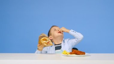 Çocuk masaya oturur, fast food yer ve mavi arka planda sevinir. Sağlıklı ve sağlıksız gıda kavramı. Fast food