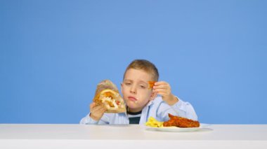 Çocuk masaya oturur, fast food yer ve mavi arka planda sevinir. Sağlıklı ve sağlıksız gıda kavramı. Fast food