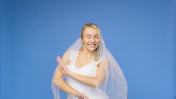 Mladá krásná blondýna se usmívá v svatební bílé šaty a závoj na modrém pozadí. Dívka se podívá do kamery. Svatba — Stock video