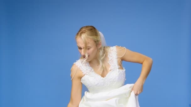 Gemarteld jonge mooie blonde vrouw in een bruiloft witte jurk en sluier op een blauwe achtergrond, doet haar schoenen uit. Het meisje kijkt in de camera. Bruiloft — Stockvideo