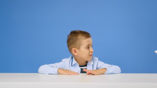 男の子の子供はテーブルに座って、青い背景にお粥を食べたくありません。悲しいと不満顔。健康的な朝食のコンセプト — ストック動画