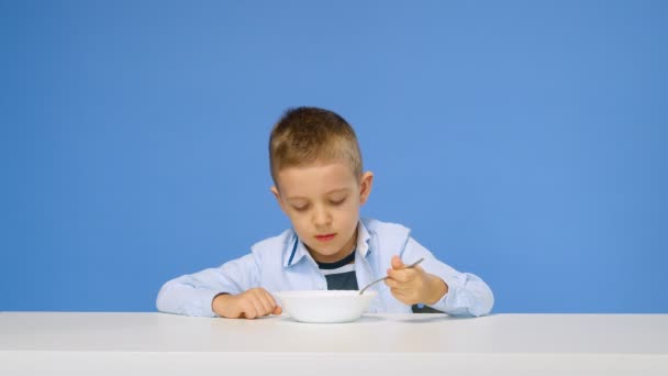 Ένα αγόρι κάθεται στο τραπέζι και δεν θέλει να φάει χυλό, σε μπλε φόντο. Λυπημένο και δυσαρεστημένο πρόσωπο. Υγιές πρωινό έννοια — Αρχείο Βίντεο