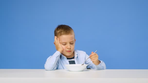 Ein Knabenkind sitzt am Tisch und will vor blauem Hintergrund keinen Haferbrei essen. Trauriges und unzufriedenes Gesicht. Konzept für gesundes Frühstück — Stockvideo