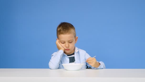 소년은 식탁에 앉아 있는데, 푸른 배경 위에서 죽을 먹고 싶어 하지 않는다. 소년은 햄버거를 받고 기뻐 합니다. 건강하고 건강하지 못한 음식의 개념. 패스트푸드 — 비디오