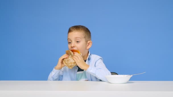 O menino está sentado à mesa e não quer comer mingau de cereal em um fundo azul. O menino é dado um hambúrguer, e ele se alegra. O conceito de comida saudável e insalubre. Fast food — Vídeo de Stock