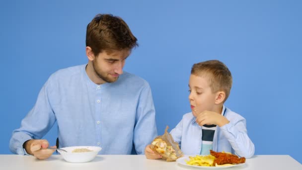 Un hombre y un niño están sentados en la mesa, el hombre está comiendo gachas, y el niño está comiendo comida rápida. El concepto de comida saludable y poco saludable. Comida rápida — Vídeo de stock