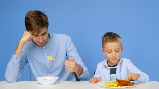 Ein Mann und ein Junge sitzen am Tisch, der Mann isst Brei, und der Junge isst Fast Food. Das Konzept der gesunden und ungesunden Ernährung. Fast food — Stockvideo