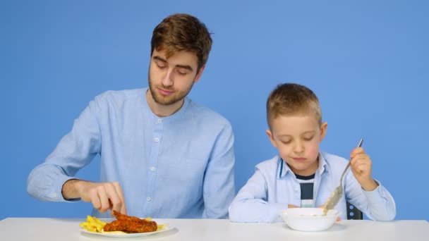 Мужчина и мальчик сидят за столом, мужчина ест фаст-фуд, а мальчик ест кашу. Понятие здоровой и нездоровой пищи. Фаст-фуд — стоковое видео