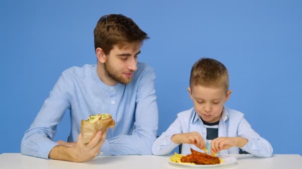 若い男と少年はテーブルに座っている、男は男の子がファーストフードを食べることを許可していません。健康的で不健康な食べ物の概念。ファーストフード — ストック動画