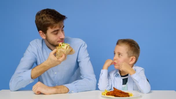 Een jongeman en een jongen zitten aan tafel, de man staat niet toe dat de jongen fastfood eet. Het concept van gezond en ongezond voedsel. Fast food — Stockvideo