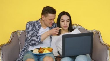 Bir erkek ve bir kız fast food yer ve sarı arka planda bir dizüstü bilgisayara bakar. Sağlıklı ve sağlıksız gıda kavramı. Fast food